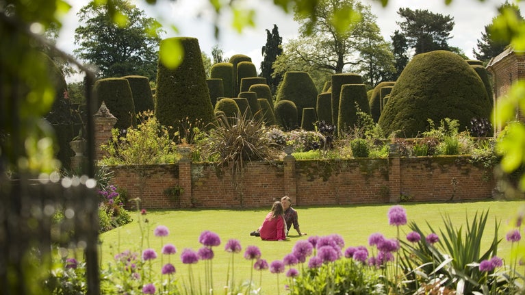 A couple sitting facing each other on the lawn in the garden at Packwood House, Warwickshire, with the yew garden in the background